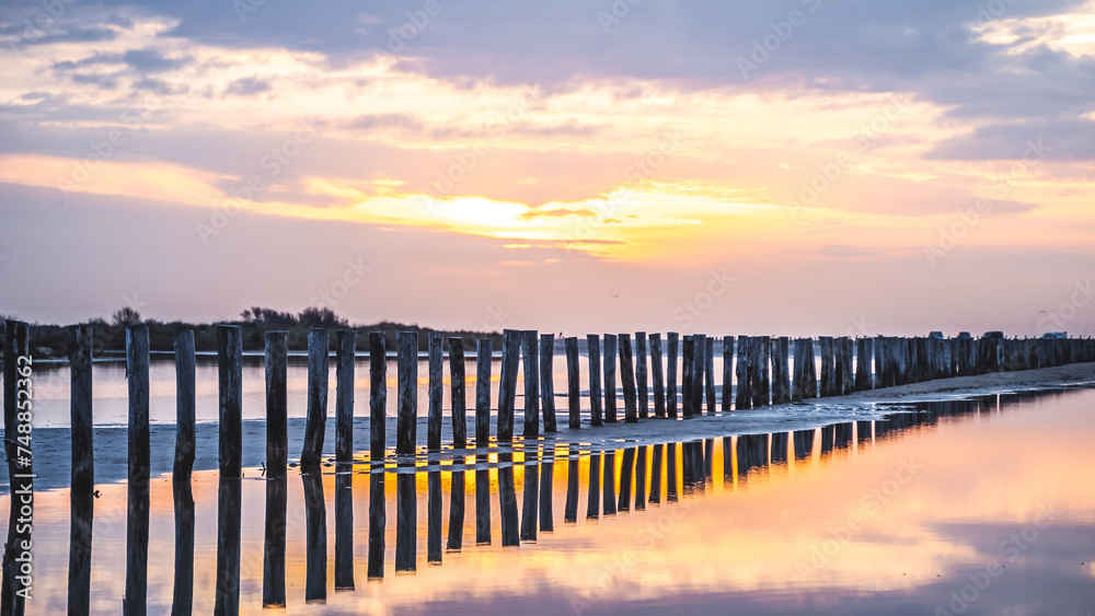 Lever du soleil et reflet dans l'eau de mer sur une plage en camargue avec des pêcheurs au loin et des poteaux en bois