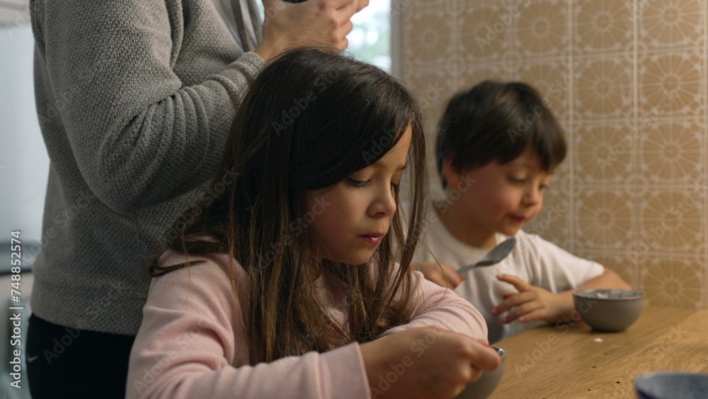 Pensive little girl eating yogurt from bowl in kitchen with mother and little brother in the background. Domestic authentic lifestyle scene of family eating breakfast early in the morning Pensive little girl eating yogurt from bowl in kitchen with mother and little brother in the background. Domestic authentic lifestyle scene of family eating breakfast early in the morning