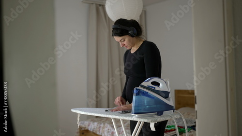 Pregnant woman ironing clothes at homedoing domestic chores while wearing headphones listening to content online, person multi-tasking photo