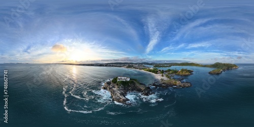 360 aerial photo taken with drone of waves crashing against rocks on island with a fort and sunset behind