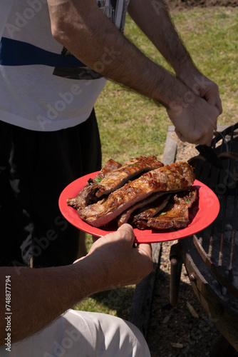 Barbecue. Caucasian male hands serving sliced roasted pork meat in a dish, besides the outdoor grill.	
 photo