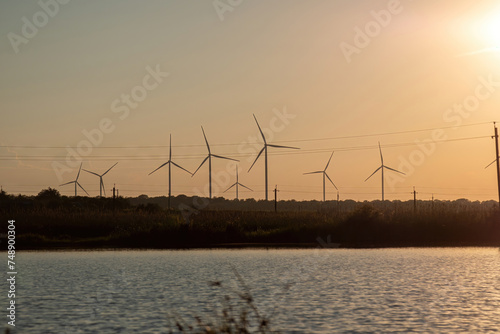 Windmills with rotor blades generating ecological energy on river bank at twilight. Production of renewable source of energy