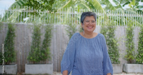 An elegant senior woman with glasses stands confidently in a garden, her wisdom and contentment reflected in her gentle smile, surrounded by lush foliage