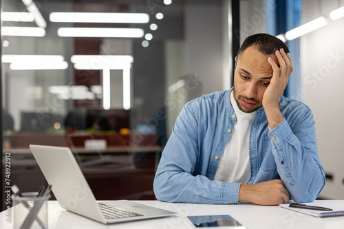 Upset and tired young hispanic man working in a cluttered office sitting sullenly at a desk with a laptop, thoughtfully resting his head on his hand photo