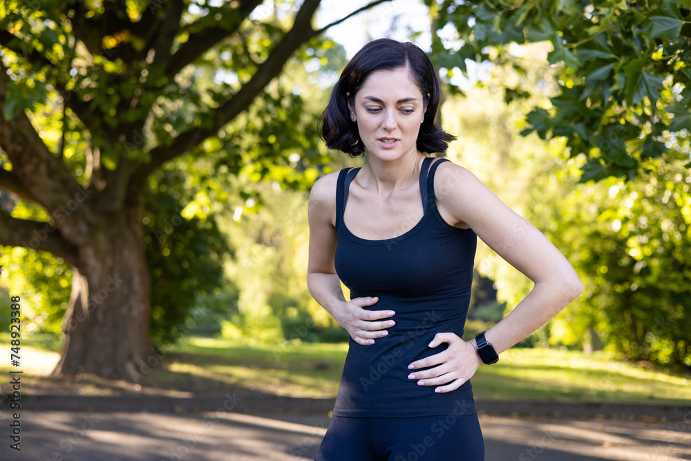 A young woman is running and doing sports in the park, standing with her hands on her stomach, feeling nauseous and near after exercise A young woman is running and doing sports in the park, standing with her hands on her stomach, feeling nauseous and near after exercise