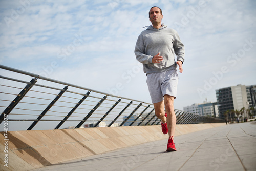 Handsome sportsman, athletic man running on the bridge on sunny day, enjoying his morning jog on the promenade