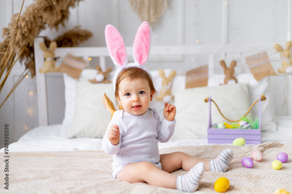 Fototapeta premium cute happy baby girl in bunny ears on the bed at home with a rabbit and painted eggs, easter concept, funny Easter baby is waiting for the holiday and rejoicing