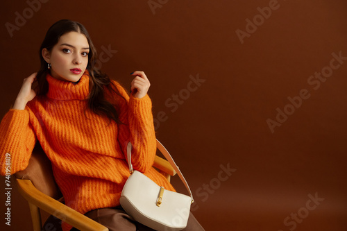 Fashionable confident woman wearing orange knitted turtleneck sweater, holding trendy white leather bag, posing on brown background. Studio fashion portrait. Copy, empty, blank space for text
