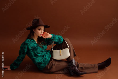 Fashionable confident woman wearing hat, green sweater, classic trousers, ankle boots, with white leather bag, posing on brown background. Studio fashion portrait. Copy, empty, blank space for text
