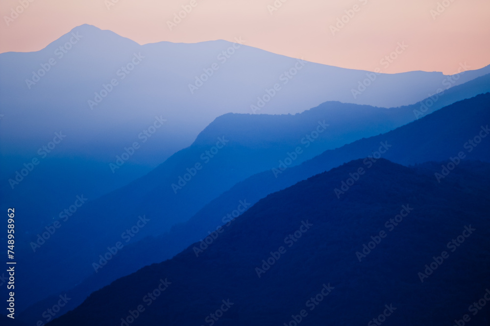 Stacked silhouettes of distant blue mountains underneath a homogenous orange evening sky at Lago Maggiore, Italy close to Switzerland. Original image with minimalist appearance.