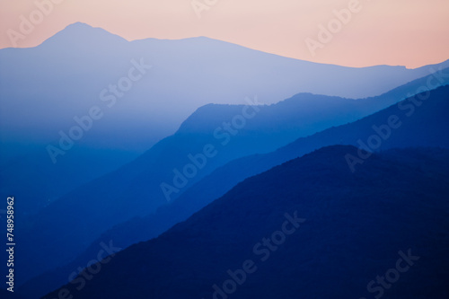 Stacked silhouettes of distant blue mountains underneath a homogenous orange evening sky at Lago Maggiore, Italy close to Switzerland. Original image with minimalist appearance.