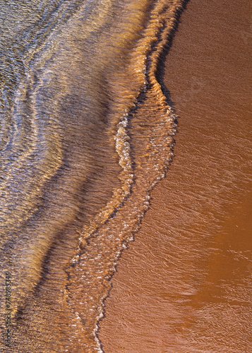 Ocean surf on sandy beach, top view. Nature background with red sand and waved lines of seawater on a sand shoreline.