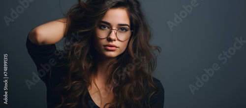 A young woman with long hair and glasses strikes a pose in front of a gray background for a photograph. She exudes confidence and style.