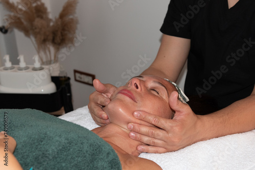 Face massage. Close up of tanned, beautiful, mature woman lying on massage table in spa salon, covered with towel, eyes closed. Facial beauty treatment. Cosmetology, Anti aging.