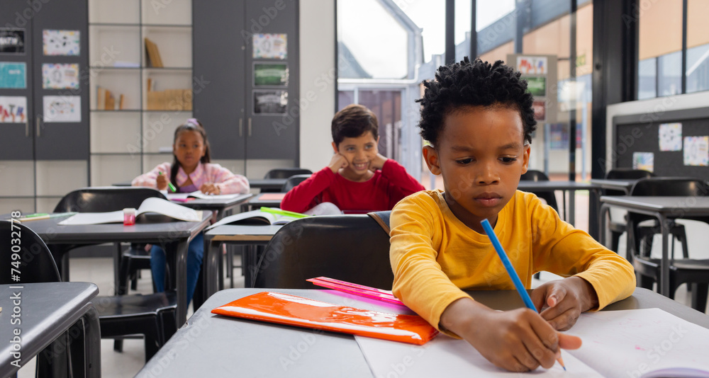 Biracial boy in yellow focuses on writing at his desk in a school classroom Biracial boy in yellow focuses on writing at his desk in a school classroom