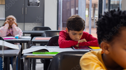Biracial boy in red looks sad at his desk in a school classroom, with copy space, classmates around  photo
