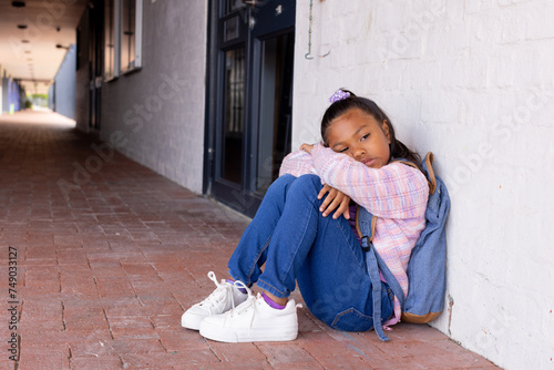 Biracial girl appears sad, sitting alone against a wall with copy space in school