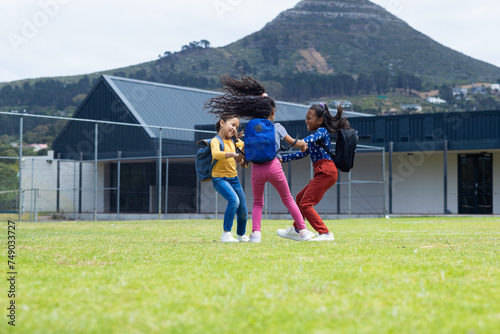 Three biracial girls are playing on a grassy field in school, with a mountain in the background
