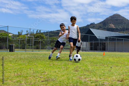 Two boys are playing soccer on a sunny day in school, with one chasing the ball
