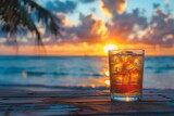 A cold glass full of a fizzy drink with ice cubes sits against a backdrop of a stunning tropical sunset at the beach