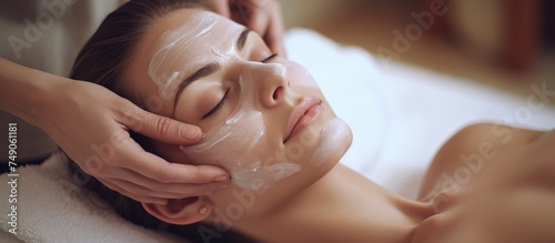 A beautiful young lady is seated with her eyes closed, receiving a facial mask treatment at a spa. A beautician is applying a thick, creamy mask to her face, ensuring even coverage for a rejuvenating