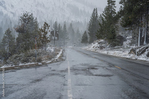 Looking downhill at wet mountain road with forest backdrop and snow during winter storm.