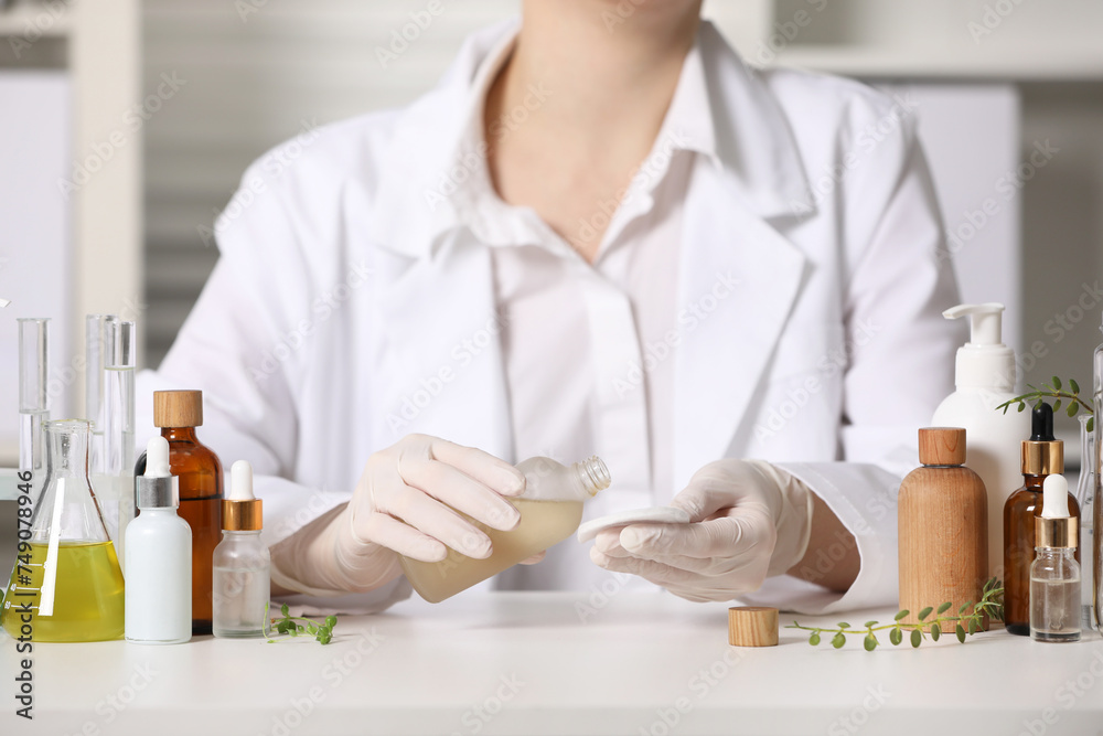 Dermatologist with bottle testing cosmetic product at white table indoors, closeup