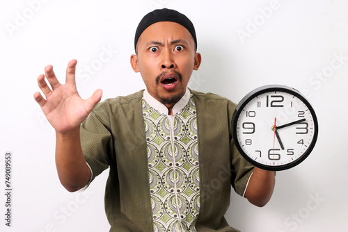 A shocked Indonesian Muslim man in koko and peci holds a clock, realizing he is late for his sahur meal during Ramadan. Isolated on a white background photo