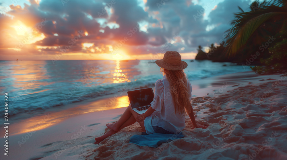 Work from anywhere. Rear view of young woman, female freelancer in straw hat working on laptop, while sitting on the tropical sandy beach at sunset