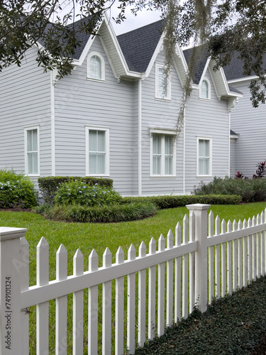 A white wooden picket fence is in the foreground with lush green grass and hedges in the garden of a large gray house. The building has multiple glass windows with white trim and a black shingled roof