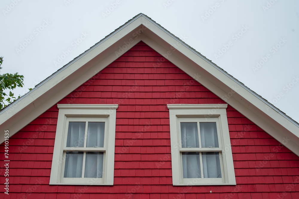 The middle roof peak of a vibrant red wooden cottage. The exterior wall of the house is covered in Cape Cod wood siding, two double-hung windows with cream trim and curtains. The sky is cloudy grey.