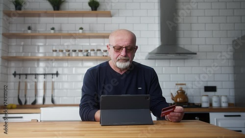 Elderly man sit in kitchen enter credit card number on tablet and due insufficient funds experiencing problems during payment online with credit card get mad photo