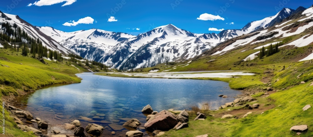 A mountain lake sits nestled among green grass and rugged rocks under the snowy peaks of Independence Pass. The tranquil water reflects the clear blue sky above.