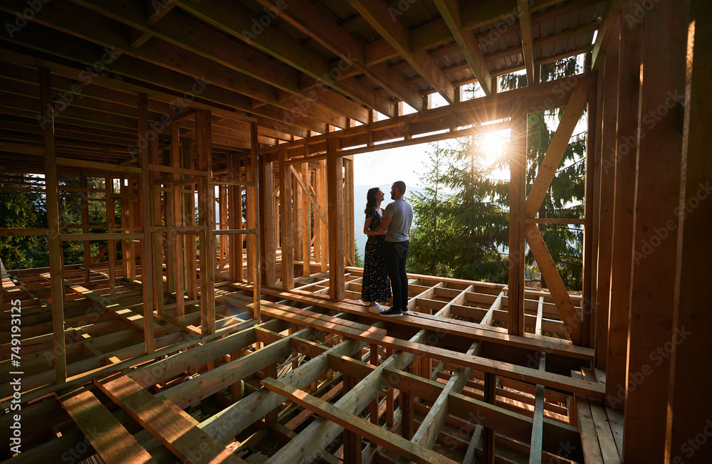 Man and woman inspecting their future wooden frame house nestled in the mountains near forest. Youthful couple at construction site in early morning. Concept of contemporary ecological construction. Man and woman inspecting their future wooden frame house nestled in the mountains near forest. Youthful couple at construction site in early morning. Concept of contemporary ecological construction.