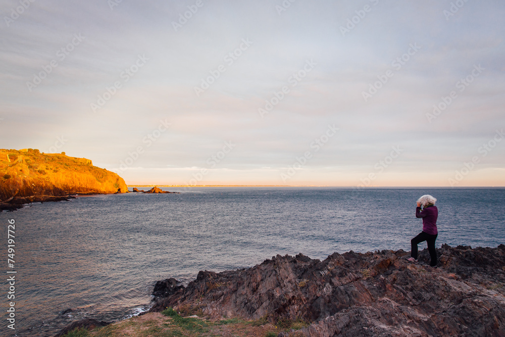 Une femme photographiant la côte méditerranée au lever de soleil. Lever de soleil sur le littoral. Touriste au bord de la mer en hiver. Lumière dorée sur la côte rocheuse.