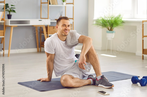 Portrait of an attractive sporty young man wearing white T-shirt sitting on the floor on yoga mat and looking cheerful at the camera after doing sport exercises at home holding a bottle of water.