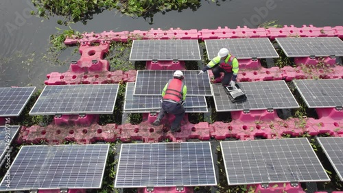 Aerial top view two technician workers use laptop and tools to check and maintenance solar cell panels over water reservoir.