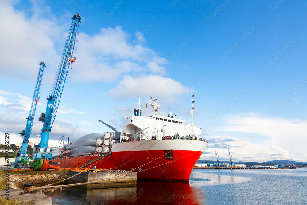 Transport ship moored to the pier of a seaport loaded with wind generator blades, with port truck cranes standing nearby.