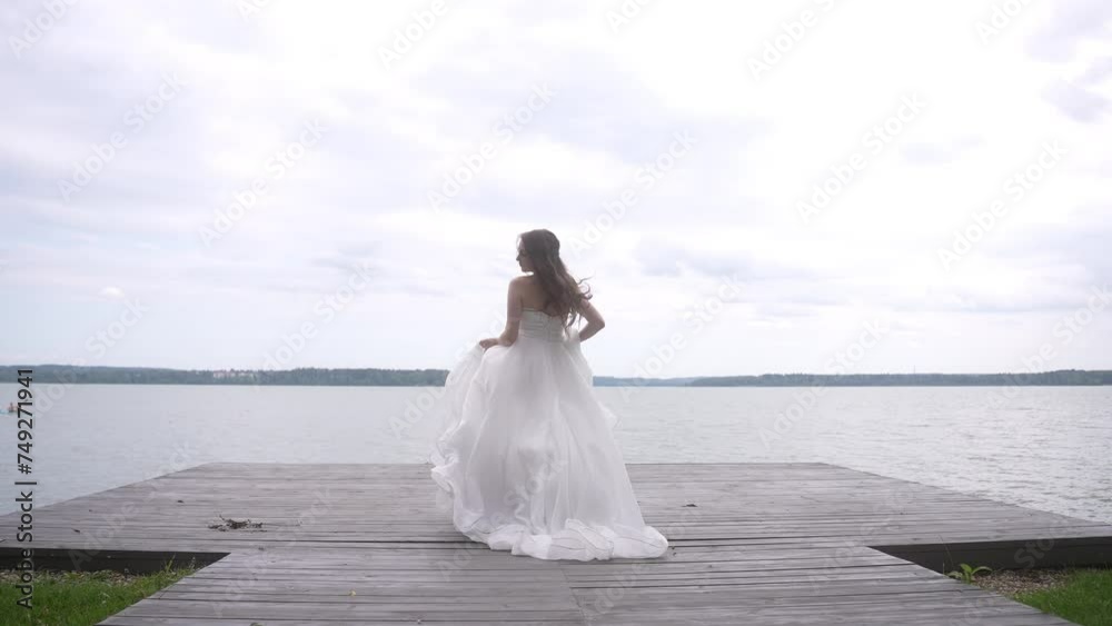 A bride stands at the lakeshore, flowers in hand, smiling under a cloudy sky A bride stands at the lakeshore, flowers in hand, smiling under a cloudy sky