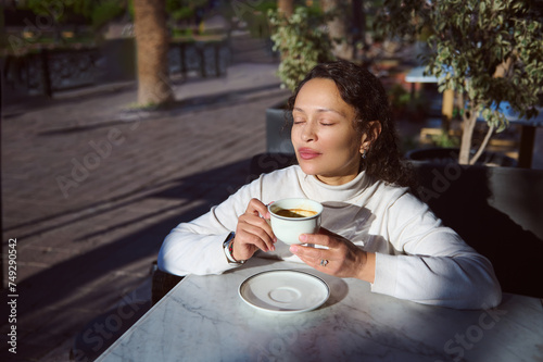 Delightful relaxed young woman sitting at table in an outdoor street bar, drinking hot tea while stanging outdoors at city street photo