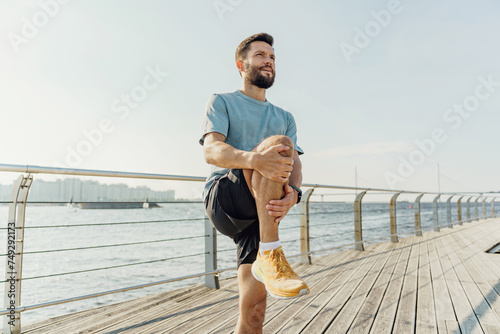Man in athletic gear doing knee stretches on a sunny boardwalk by the sea.