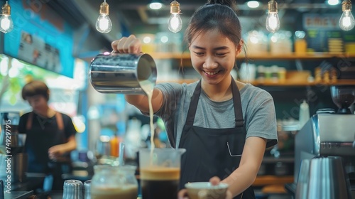 Young adult asian female woman barista pouring fresh milk to prepare latte coffee for customer in cafe bar with her colleague working in background. For small business startup in food industry concept photo