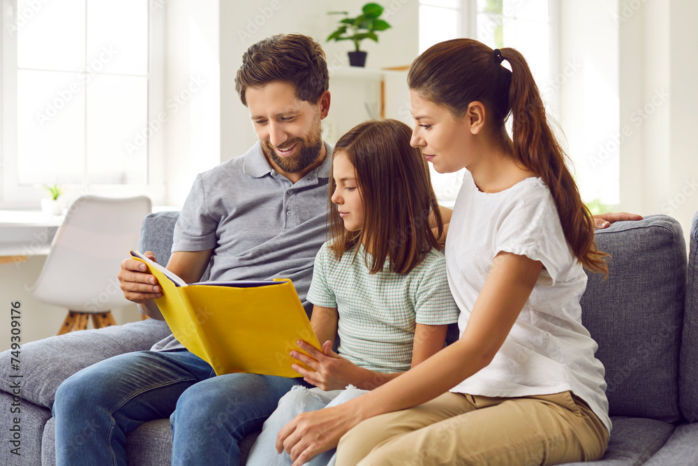 Portrait of a young happy family reading a book with their daughter sitting on sofa at home. Parents spending time with their child and smiling. Family leisure time together concept.