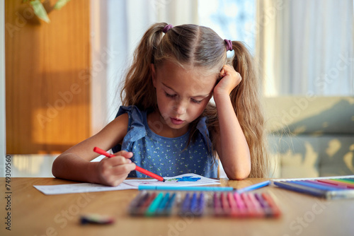 Focused little schoolgirl with ponytails in blue dress sitting at wooden table leaning on hand and painting picture with multicolored felt tip pens during weekend at home on sunny day