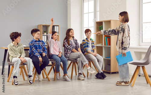 Group of smiling school children in casual wear sitting in row in classroom. Schoolgirl acting as teacher and asking question to her classmates. School education  discussion