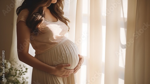 Young attractive pregnant mother in beige outfit staying at the home window , touching and looking at belly with happy expression. Maternity concept