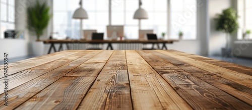 A wooden table is placed in a room flooded with natural light from numerous windows. The table is bare, with no objects on it, set against a backdrop of a blurred office or meeting room.