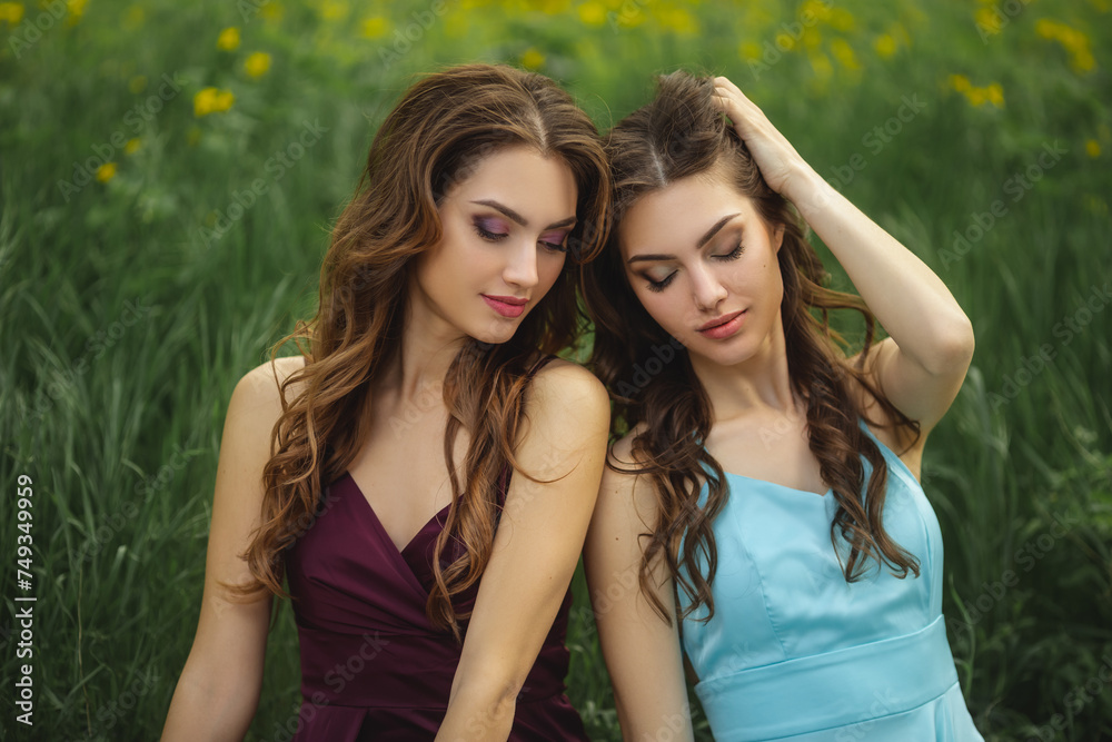 Portrait of fashionable twins models with perfect make-up and hairstyle sitting on green grass meadow with yellow flowers. Spring beauty concept. Portrait shot of two gorgeous women in evening dresses