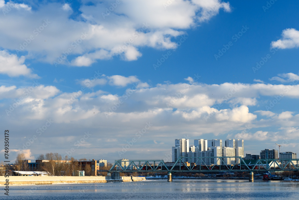 A block of new houses on the river embankment and a railway bridge against the backdrop of a high blue sky with clouds. 