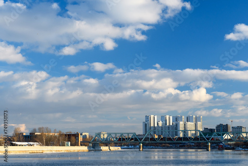 A block of new houses on the river embankment and a railway bridge against the backdrop of a high blue sky with clouds. 